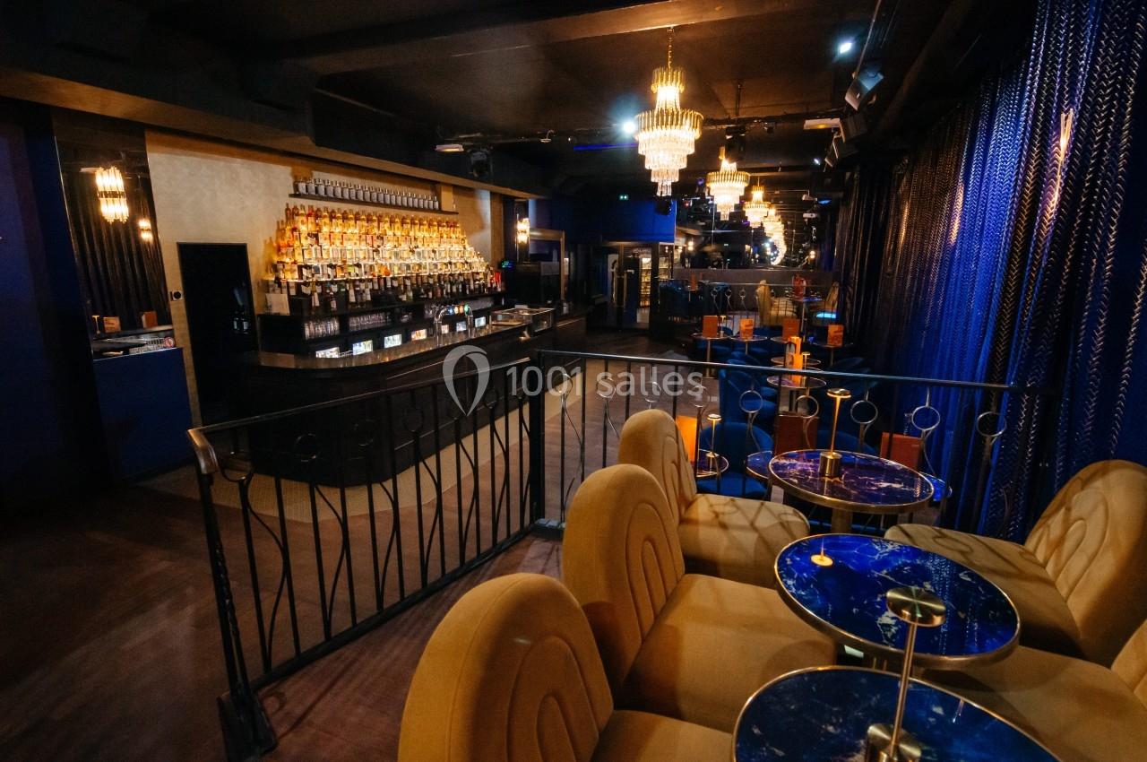 Intérieur d'un bar lounge avec comptoir éclairé, tables en marbre bleu et fauteuils rembourrés sous des lustres.