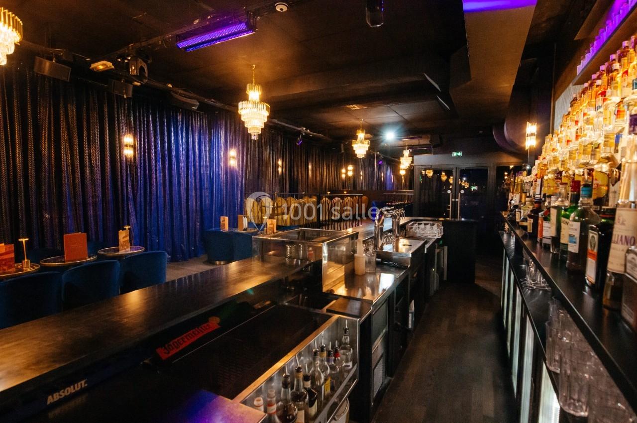 Intérieur d'un bar avec comptoir, rangées de bouteilles, tables et chaises sous un éclairage tamisé.