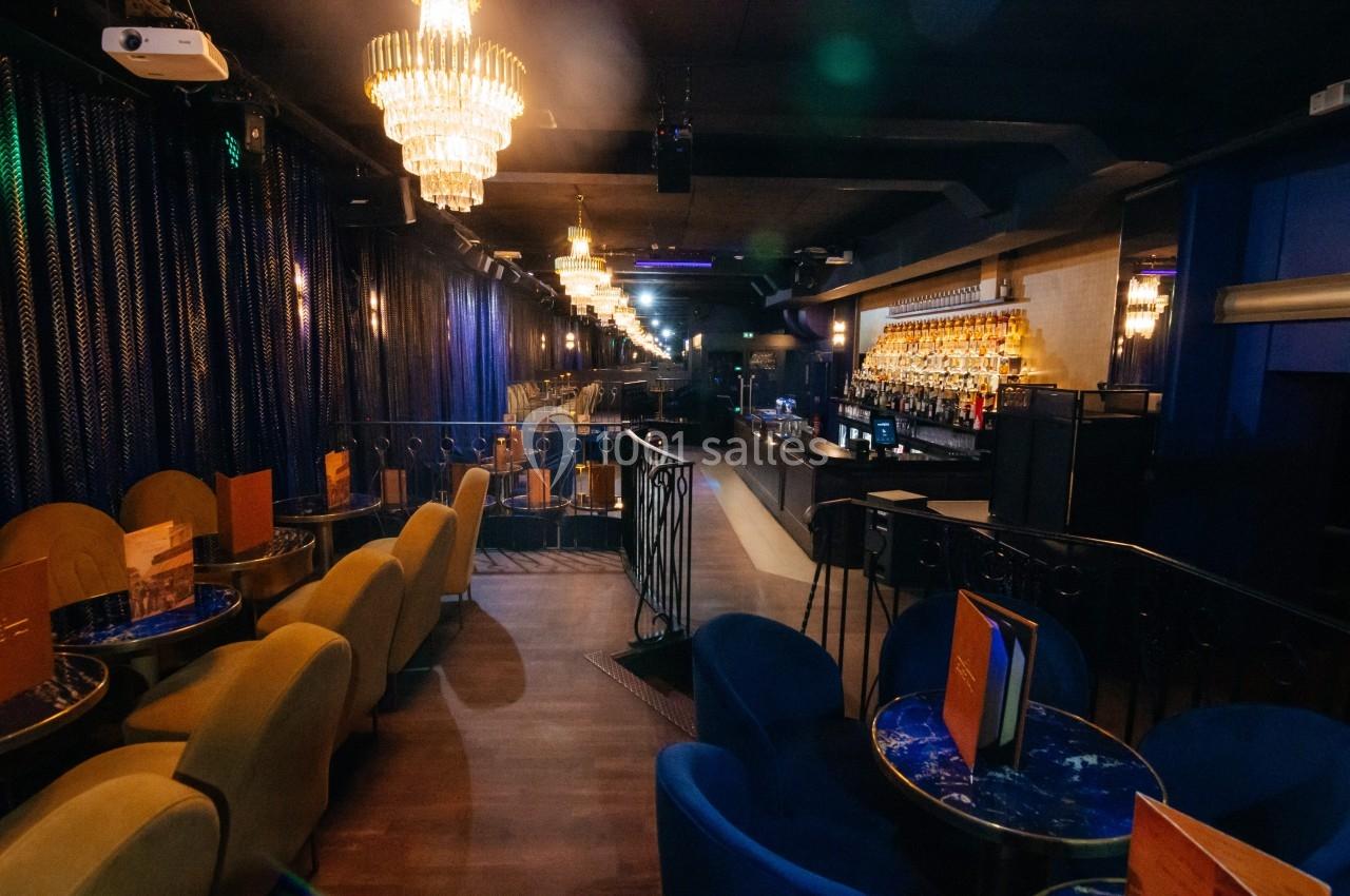 Salle de bar élégante avec lustres, fauteuils colorés, tables rondes et un comptoir illuminé en arrière-plan.