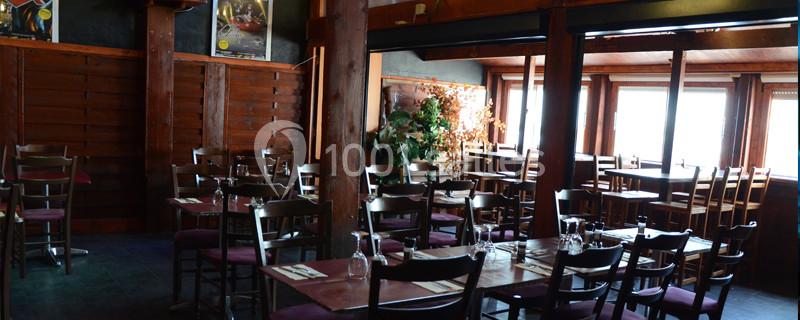 Salle de restaurant en bois avec tables dressées, chaises alignées et lumière naturelle entrant par de grandes fenêtres.