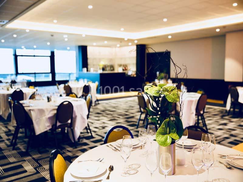 Salle de réception lumineuse avec tables rondes dressées, nappes blanches et décoration florale au centre.