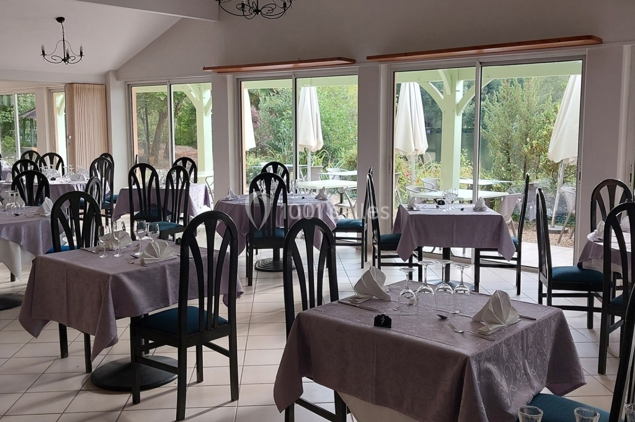 Salle de restaurant lumineuse avec tables dressées, nappes violettes et vue sur une terrasse extérieure verdoyante.