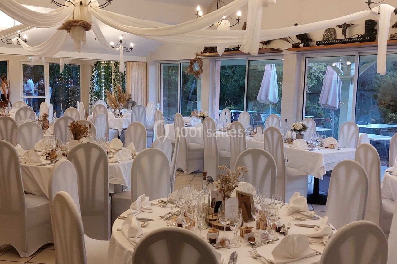 Salle de réception décorée avec des tables rondes, nappes blanches et chaises habillées, donnant sur un jardin extérieur.