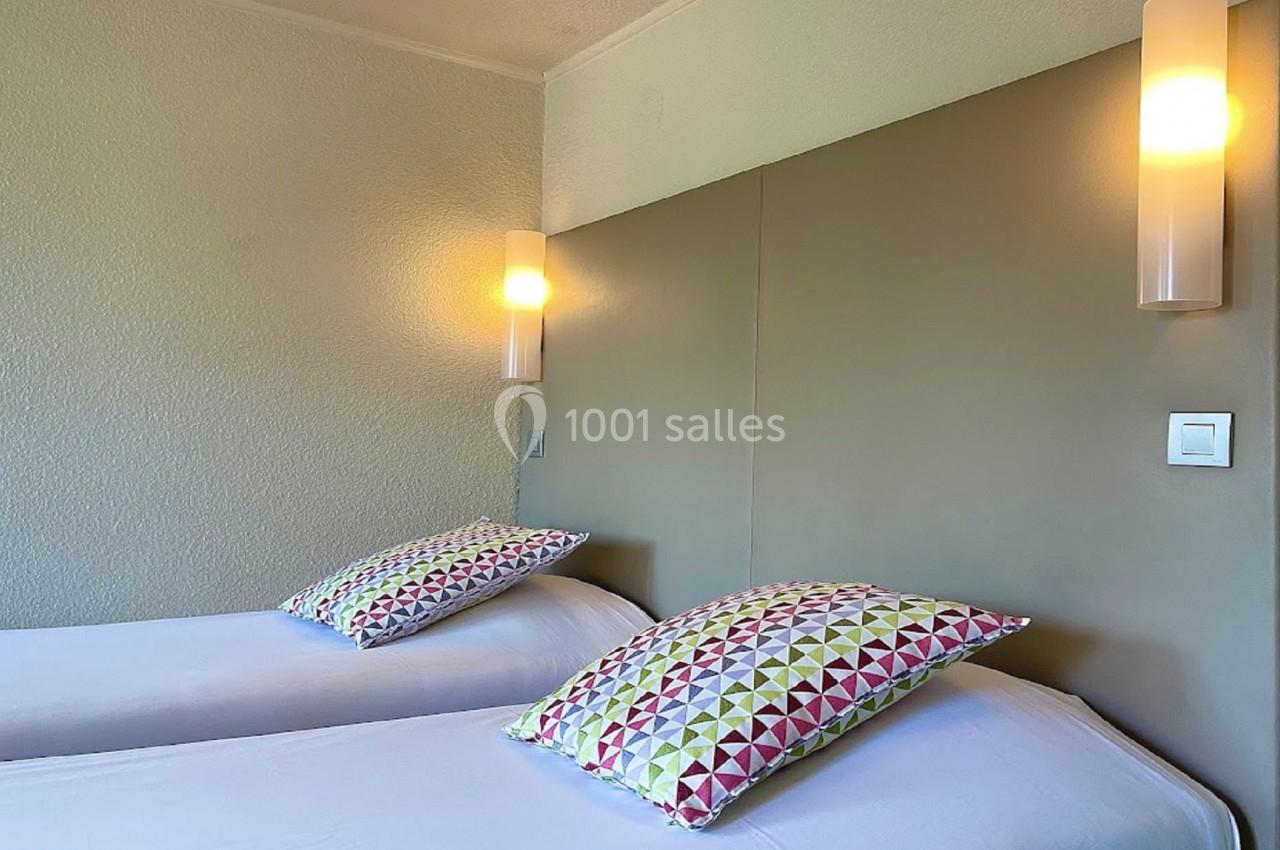 Deux lits simples avec des coussins à motifs géométriques colorés, éclairés par deux appliques murales.