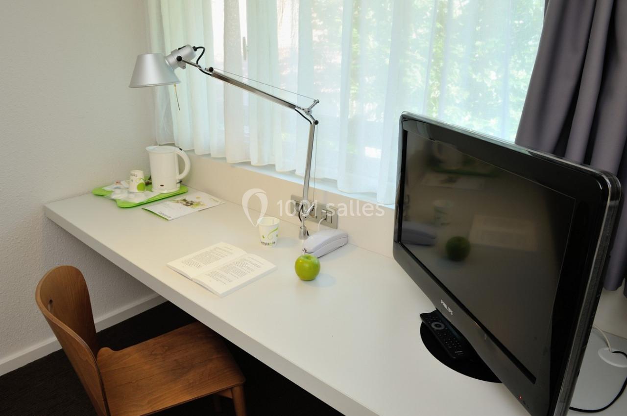 Bureau avec lampe articulée, télévision, chaise en bois, livre ouvert, pomme verte et plateau avec bouilloire.