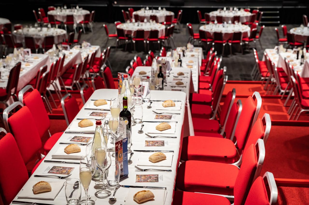 Tables dressées avec nappes blanches, verres, bouteilles et assiettes, entourées de chaises rouges dans une grande salle.