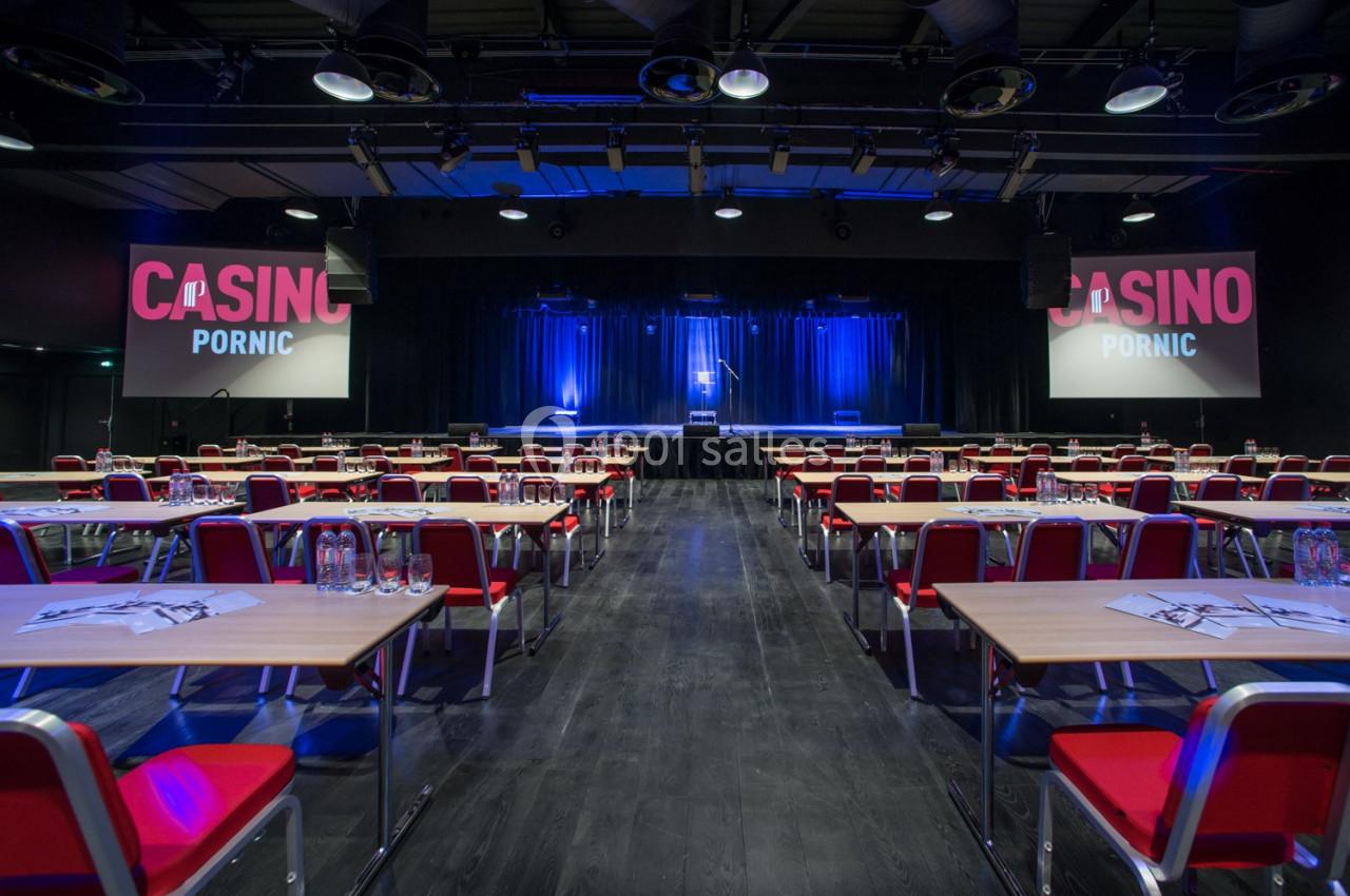 Salle de conférence avec des tables et chaises rouges, éclairage bleu sur une scène au fond, écrans affichant ’Casino Pornic…