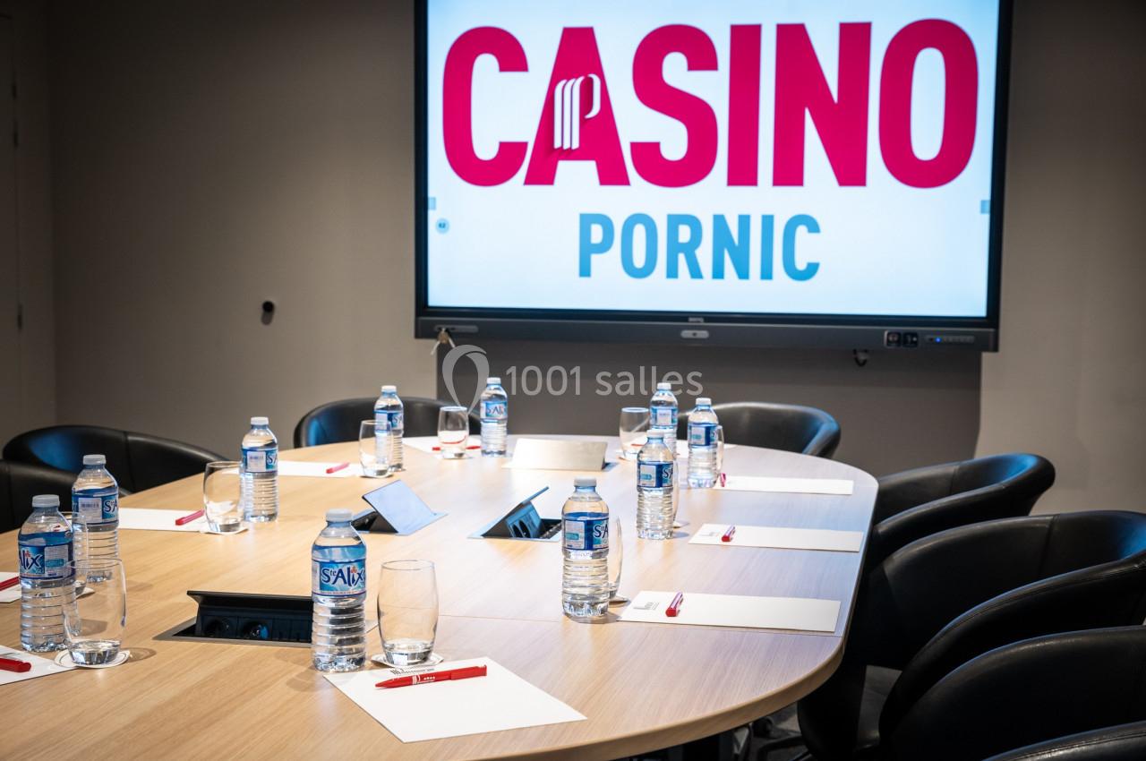 Salle de réunion avec table ovale, chaises noires, bouteilles d'eau, blocs-notes, stylos et écran affichant ’Casino Pornic’.