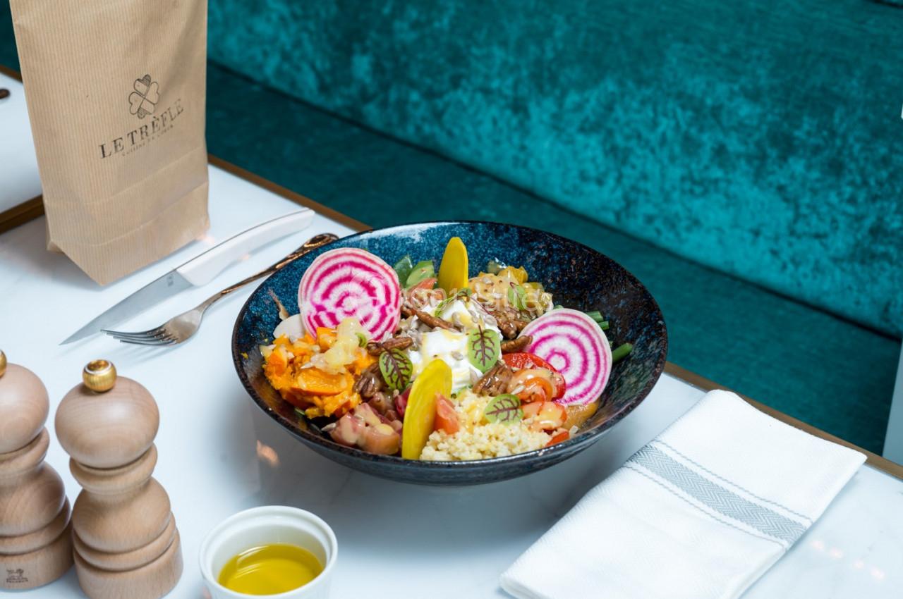 Bol de salade colorée avec légumes, betteraves, couscous et sauce, servi sur une table avec couverts et huile d'olive.