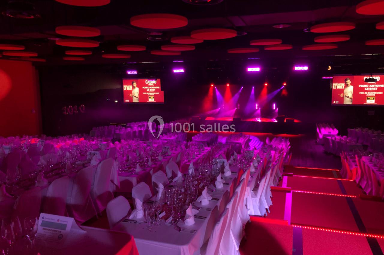 Salle de réception éclairée en rouge avec tables dressées et une scène illuminée en arrière-plan.