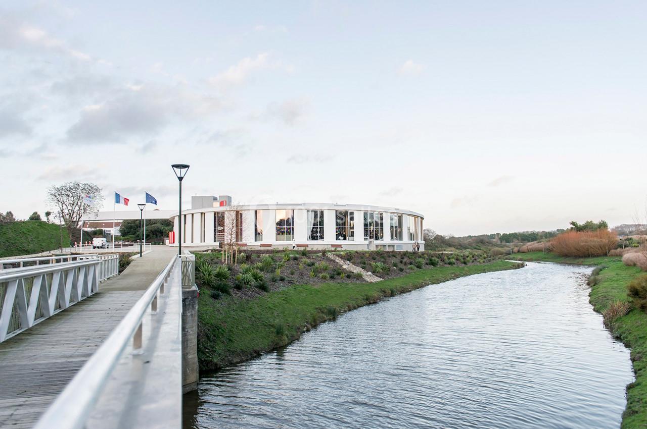 Bâtiment moderne en bord de rivière, entouré de verdure, avec un pont piétonnier au premier plan.