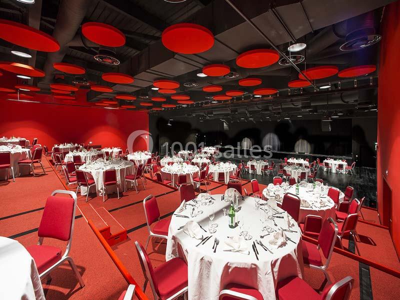Salle de réception avec tables rondes dressées, chaises rouges et plafond décoré de panneaux acoustiques rouges.