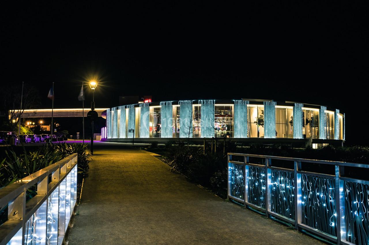 Bâtiment circulaire moderne illuminé la nuit, avec un chemin éclairé et des rampes décorées de lumières.