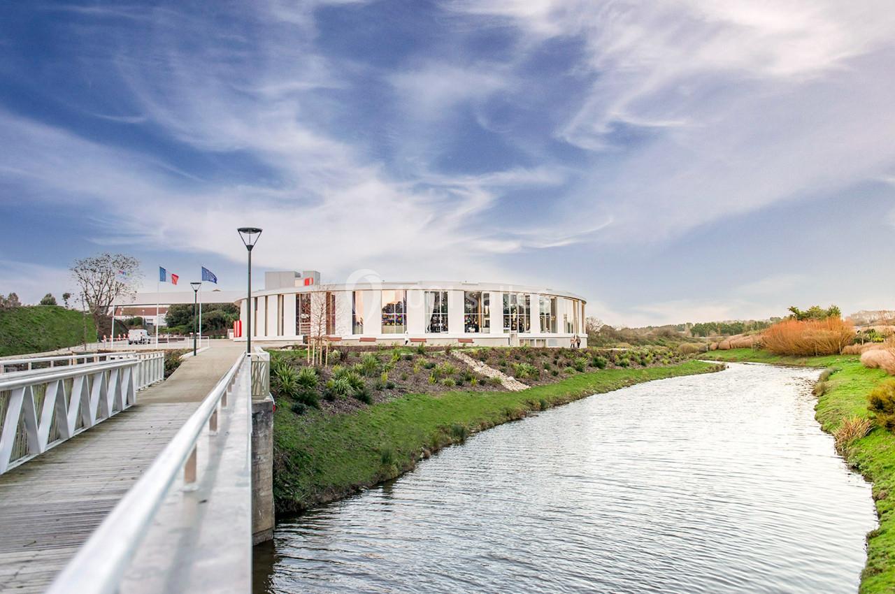 Bâtiment moderne en bord de rivière, entouré de verdure, avec un pont en bois au premier plan.
