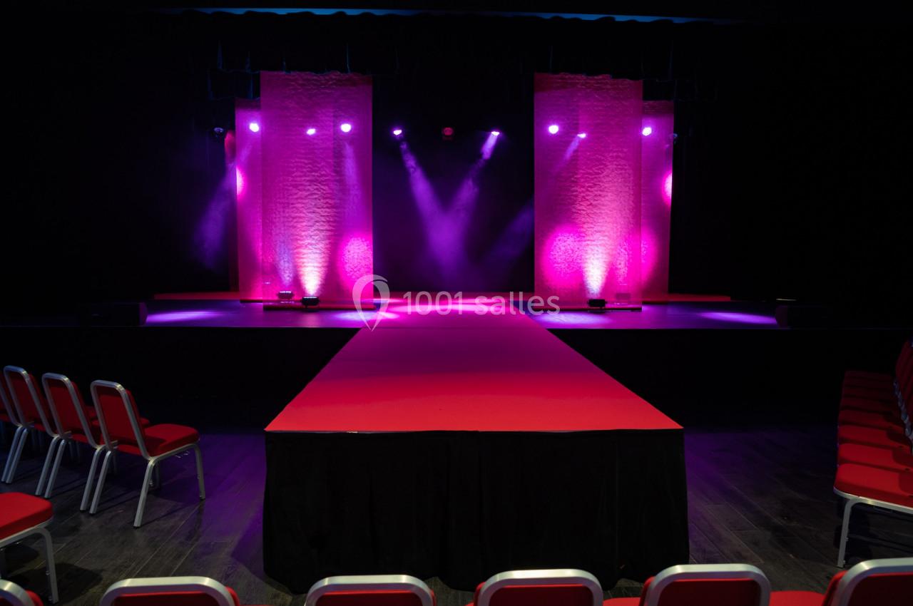 Scène avec éclairage rose et violet, podium central rouge entouré de chaises vides.