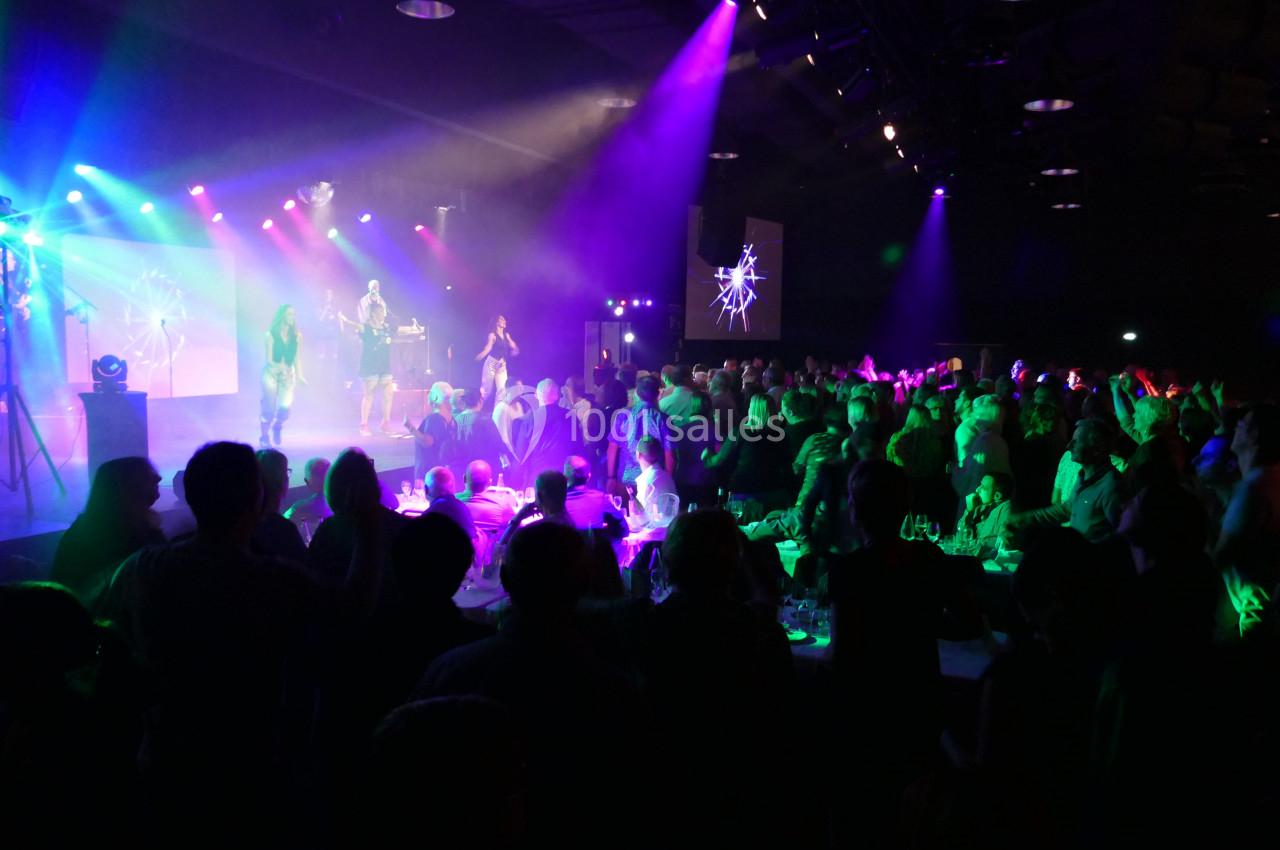 Groupe de chanteurs sur scène éclairée par des lumières colorées, devant un public nombreux dans une salle sombre.