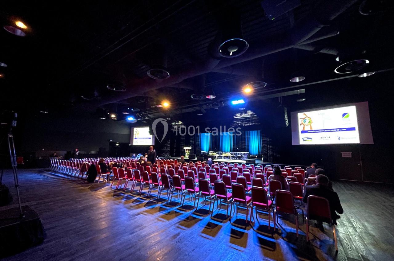 Salle de conférence sombre avec des rangées de chaises rouges, une scène éclairée et des écrans affichant des présentations.