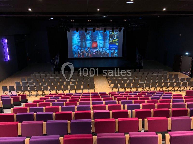 Salle de spectacle avec des rangées de sièges colorés face à une scène éclairée et un écran projetant une image.
