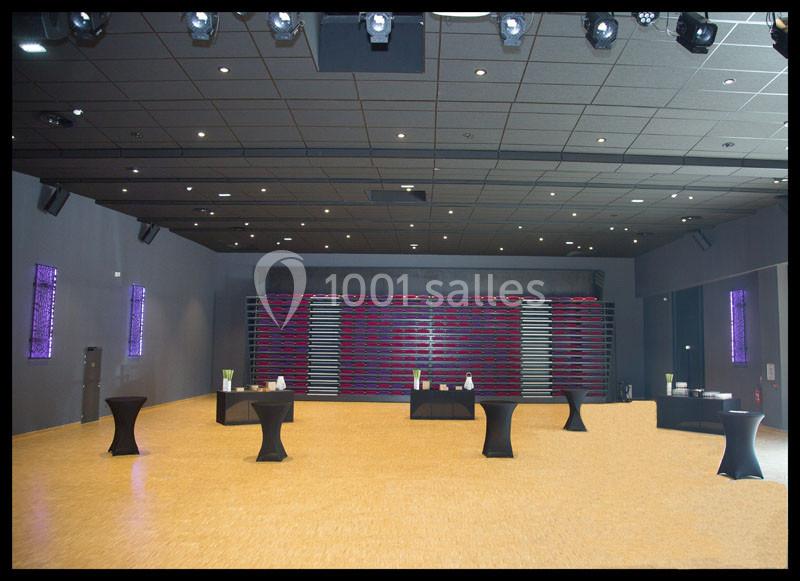 Salle vide avec parquet clair, tables hautes noires dispersées et gradins repliés au fond sous un éclairage artificiel.