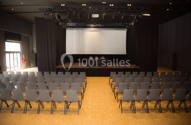 Salle de conférence avec des rangées de chaises vides face à une scène équipée d'une table et d'un écran de projection.