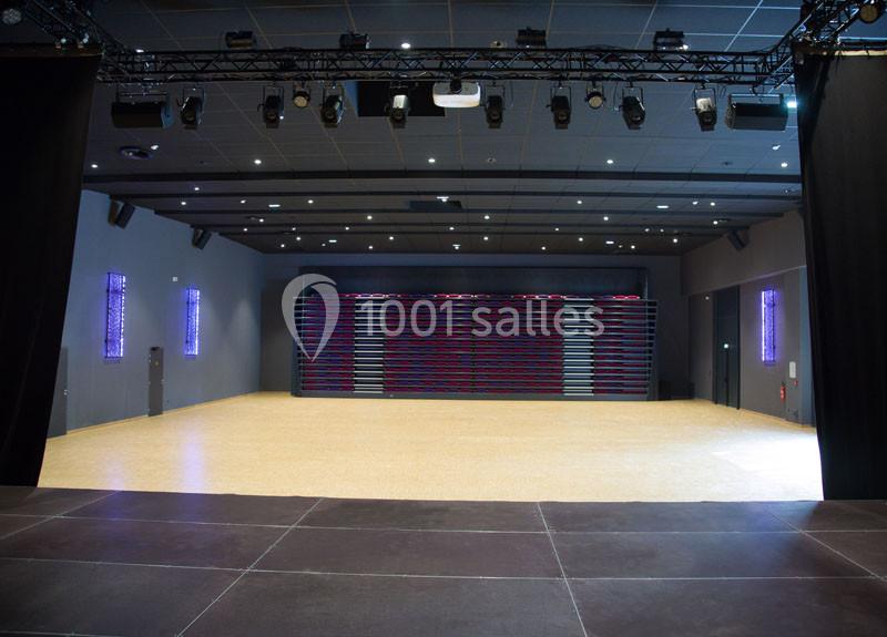 Salle polyvalente vide avec parquet clair, gradins repliés au fond, éclairage suspendu et murs sombres.