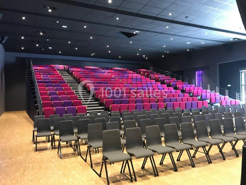 Salle de conférence avec gradins de sièges rouges et violets, et rangées de chaises noires au premier plan.