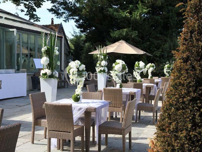 Terrasse aménagée avec des tables et chaises en rotin, décorée de compositions florales blanches, entourée de verdure.