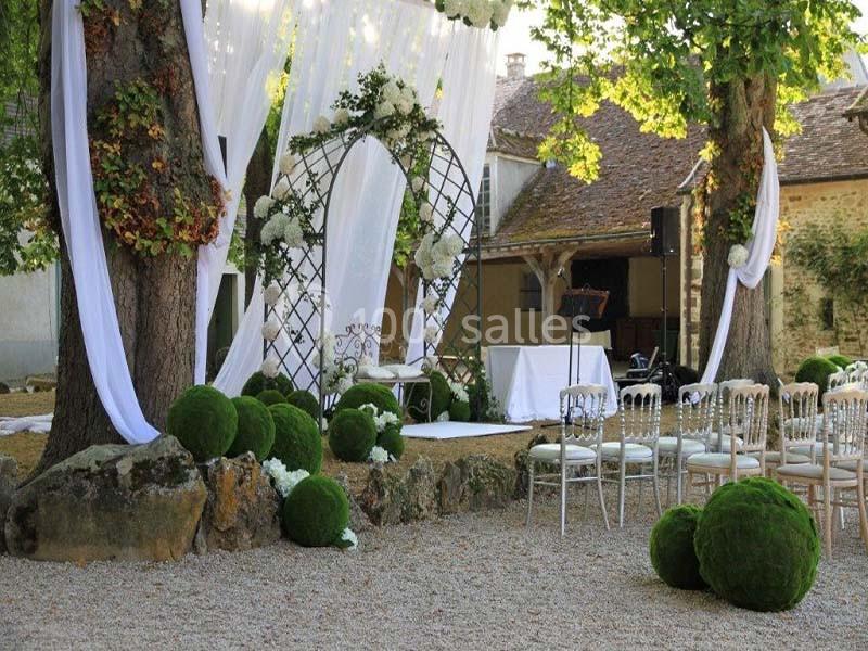 Décoration de mariage en extérieur avec arche fleurie, chaises blanches, voilages et boules de mousse verte sous des arbres.
