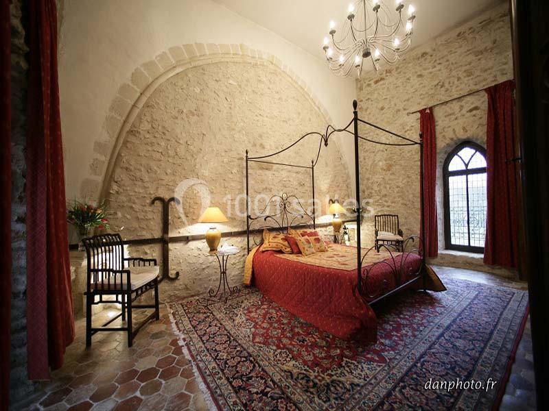 Chambre avec lit à baldaquin, murs en pierre, tapis coloré, mobilier ancien et grande fenêtre en ogive.