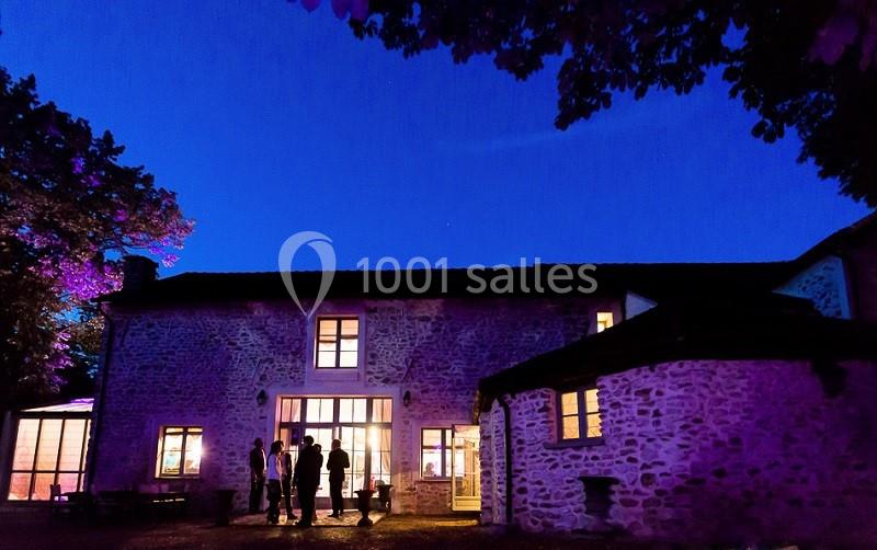 Façade d'une maison en pierre éclairée de nuit, avec des silhouettes de personnes discutant devant l'entrée.