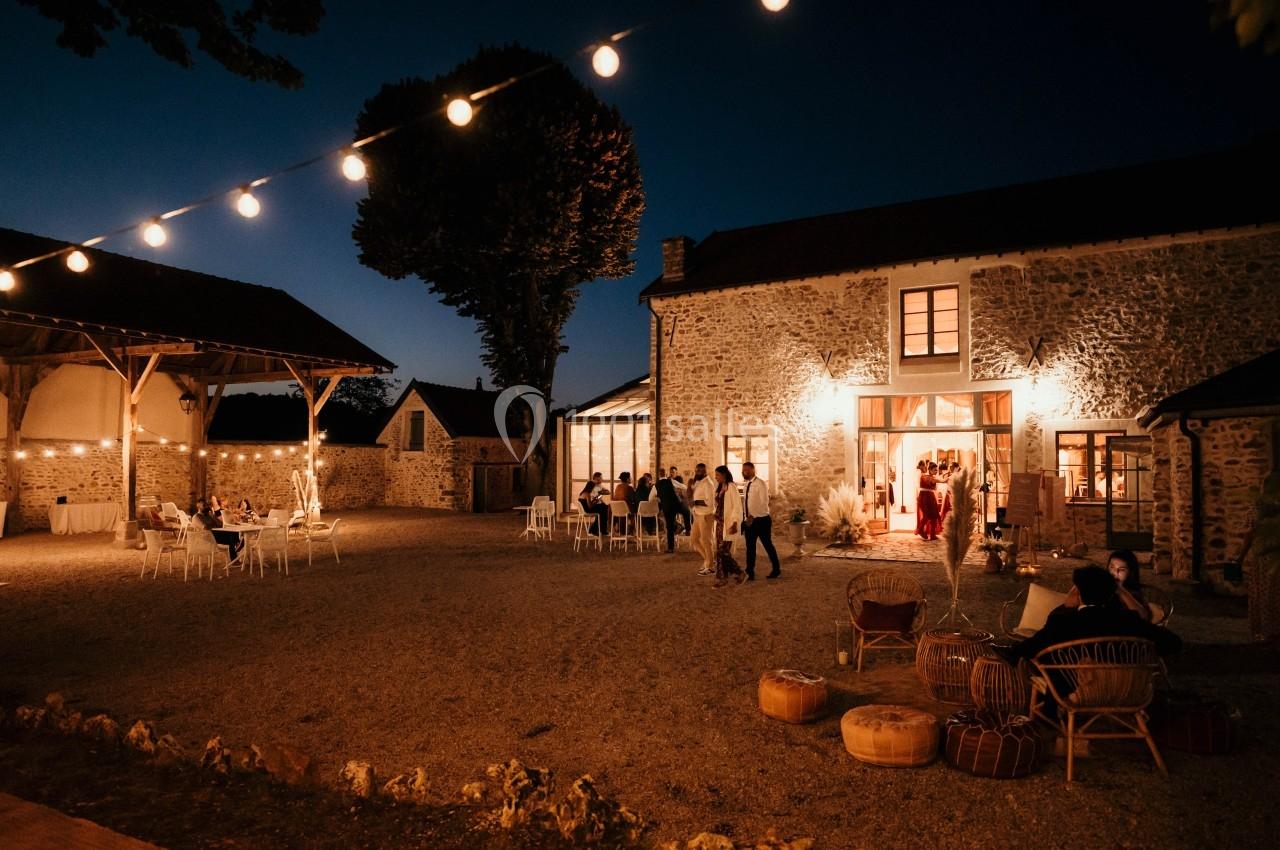 Cour d'une maison en pierre éclairée par des guirlandes lumineuses, avec des invités assis et debout en soirée.