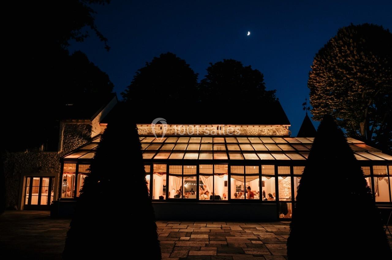 Verrière lumineuse d'une maison entourée d'arbres, éclairée de nuit sous un ciel étoilé avec une lune visible.
