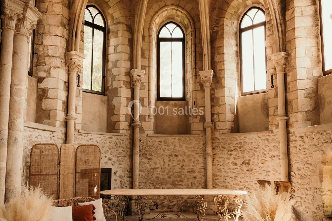 Salle voûtée en pierre avec grandes fenêtres, décorée de fleurs séchées, chaises blanches et table en bois.