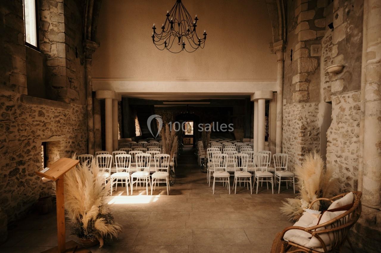 Salle en pierre avec des chaises blanches alignées, un pupitre en bois et une décoration de pampas séchées.