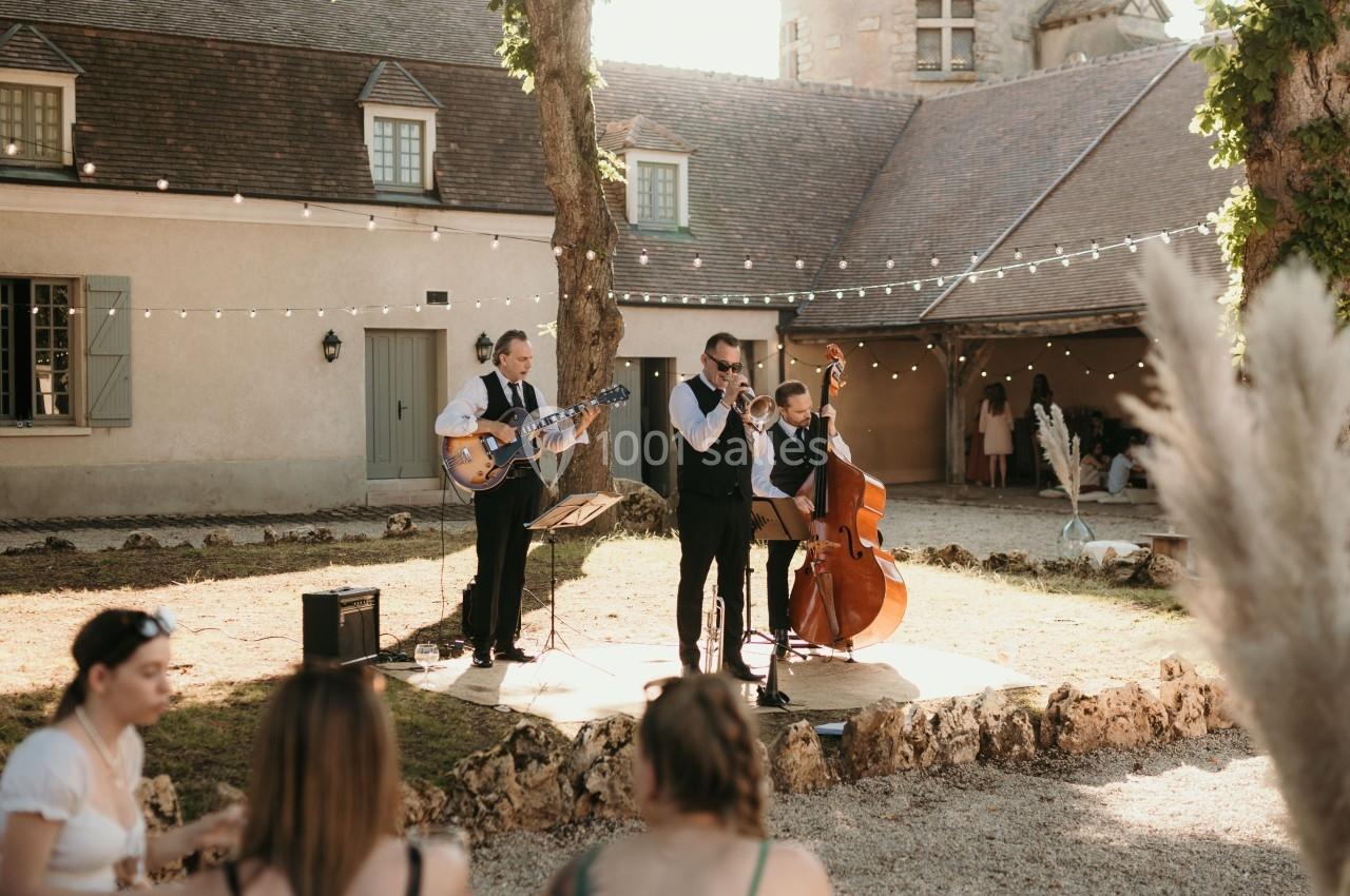 Groupe de musiciens jouant en plein air dans une cour ensoleillée, avec des invités assis au premier plan.