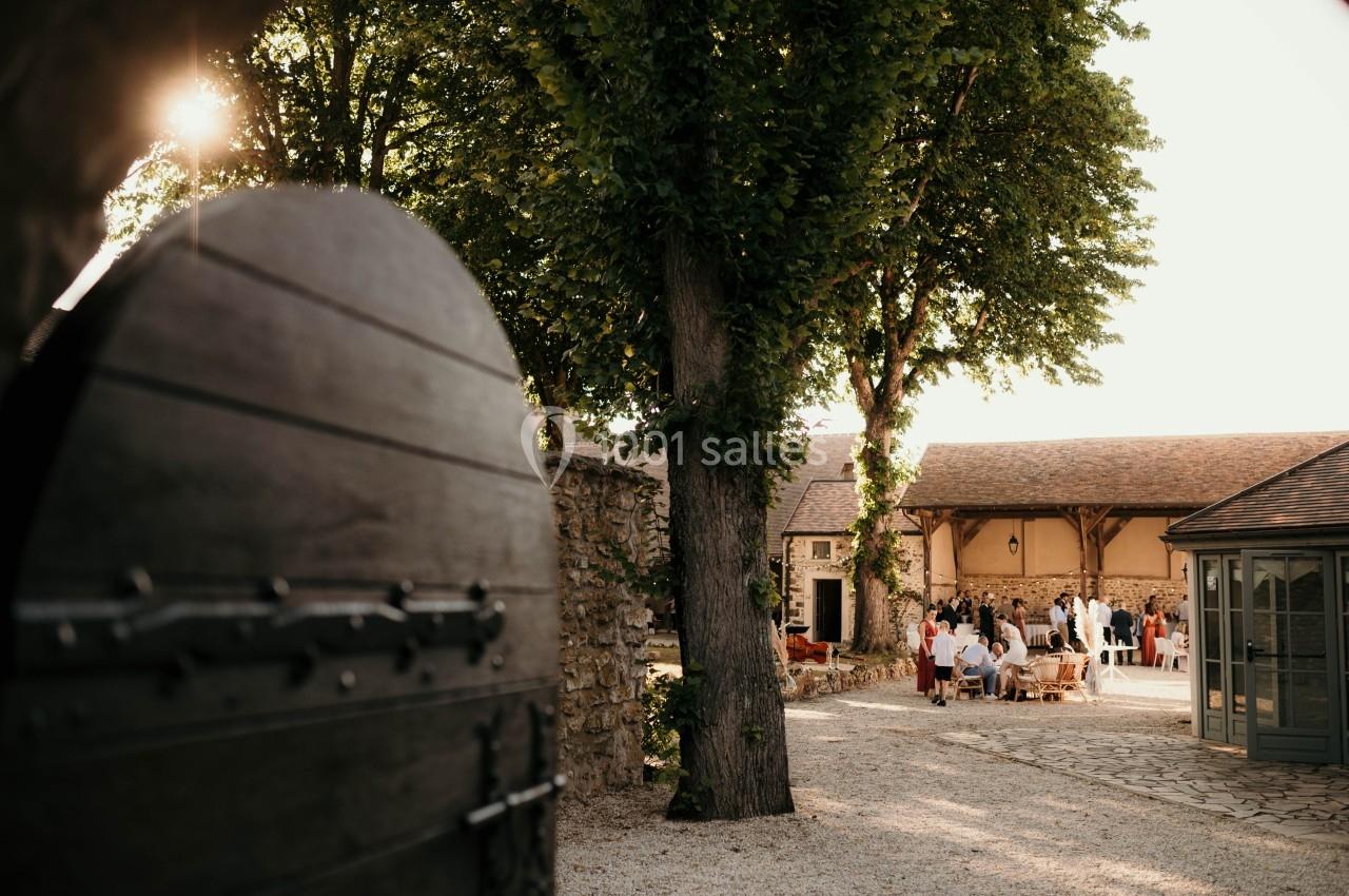 Cour intérieure ensoleillée avec des invités rassemblés près d'un bâtiment en pierre, vue depuis une porte en bois ouverte.
