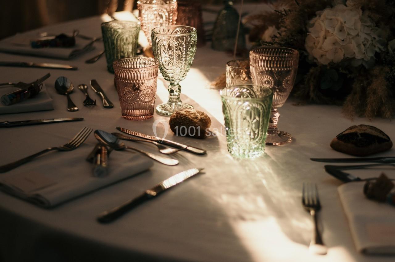 Table dressée avec nappes blanches, verres colorés, couverts et décoration florale dans une lumière tamisée.