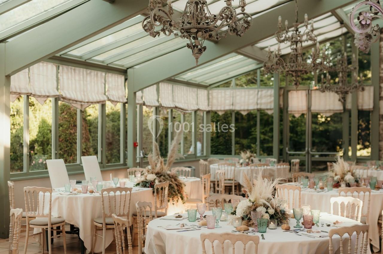 Salle lumineuse avec verrière, tables rondes dressées pour un événement, décorations florales et lustres suspendus.