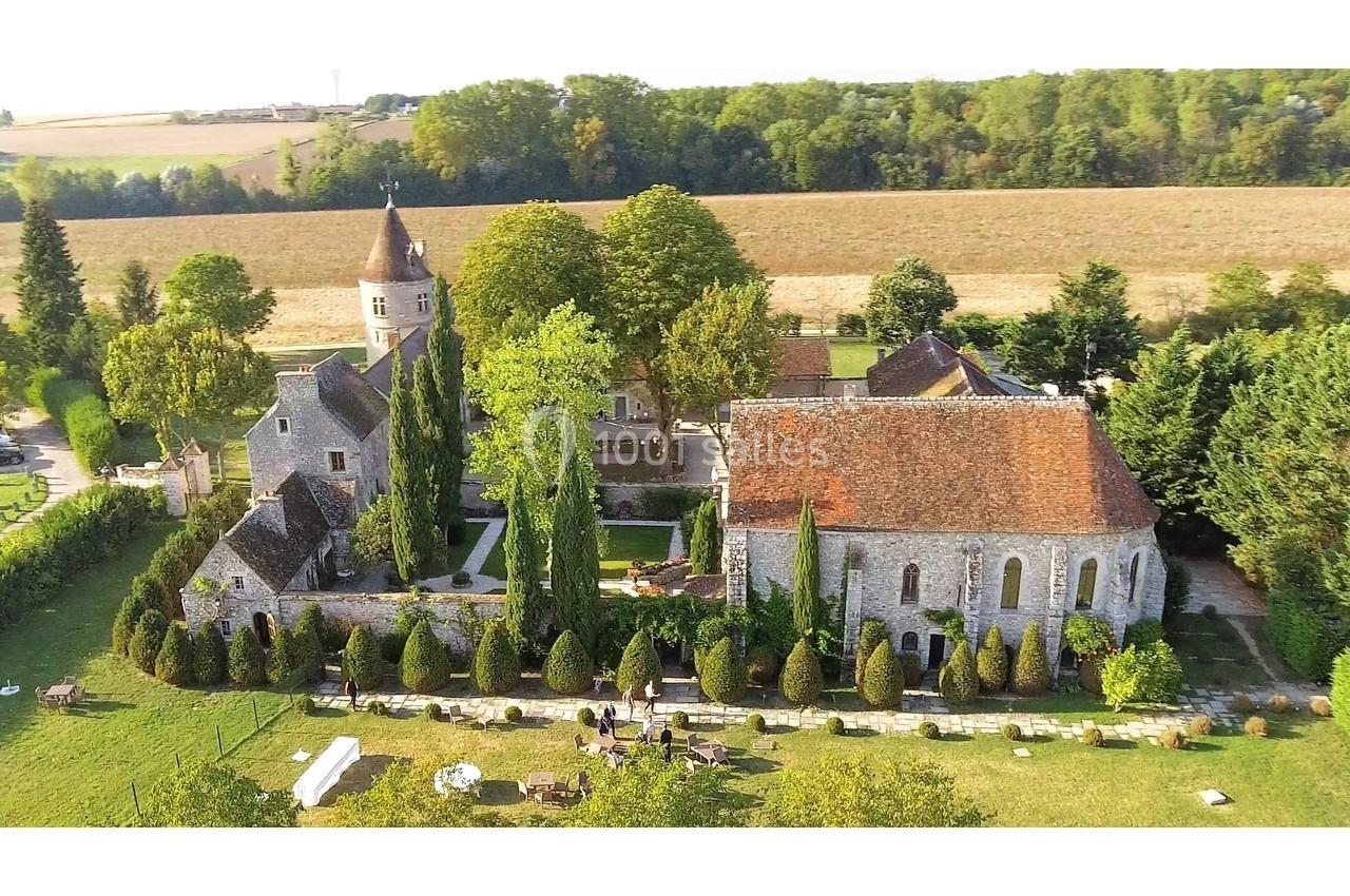 Vue aérienne d'un domaine rural avec une église en pierre, un jardin arboré et des bâtiments anciens entourés de champs.