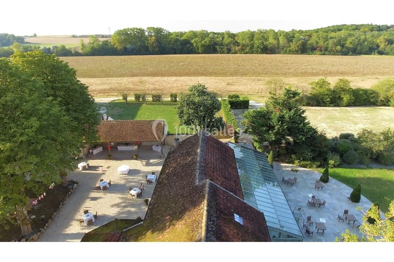 Vue aérienne d'une cour avec terrasses, bâtiments en pierre, arbres et champs en arrière-plan.