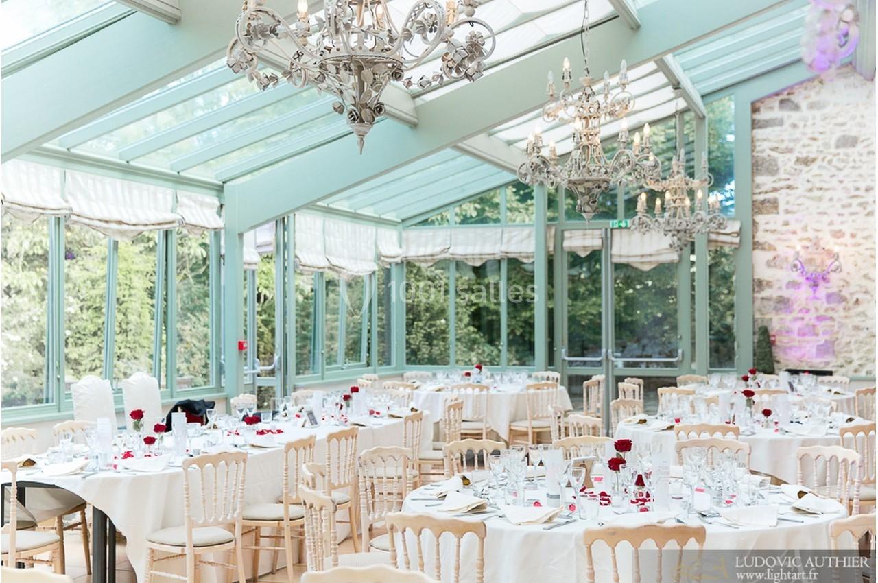 Salle de réception lumineuse avec verrière, tables dressées avec nappes blanches, chaises en bois clair et décorations…