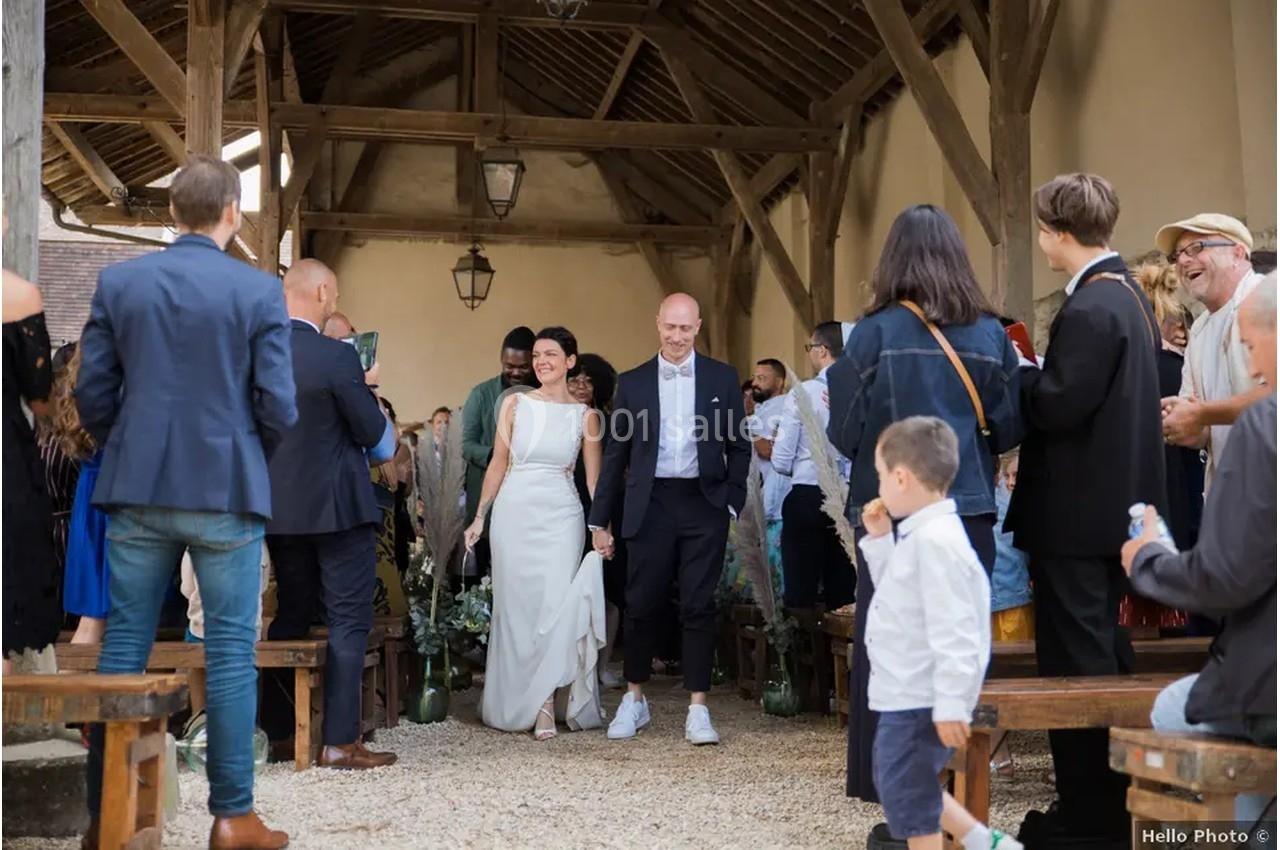 Un couple de mariés souriants marche sous une charpente en bois, entouré d'invités applaudissant.