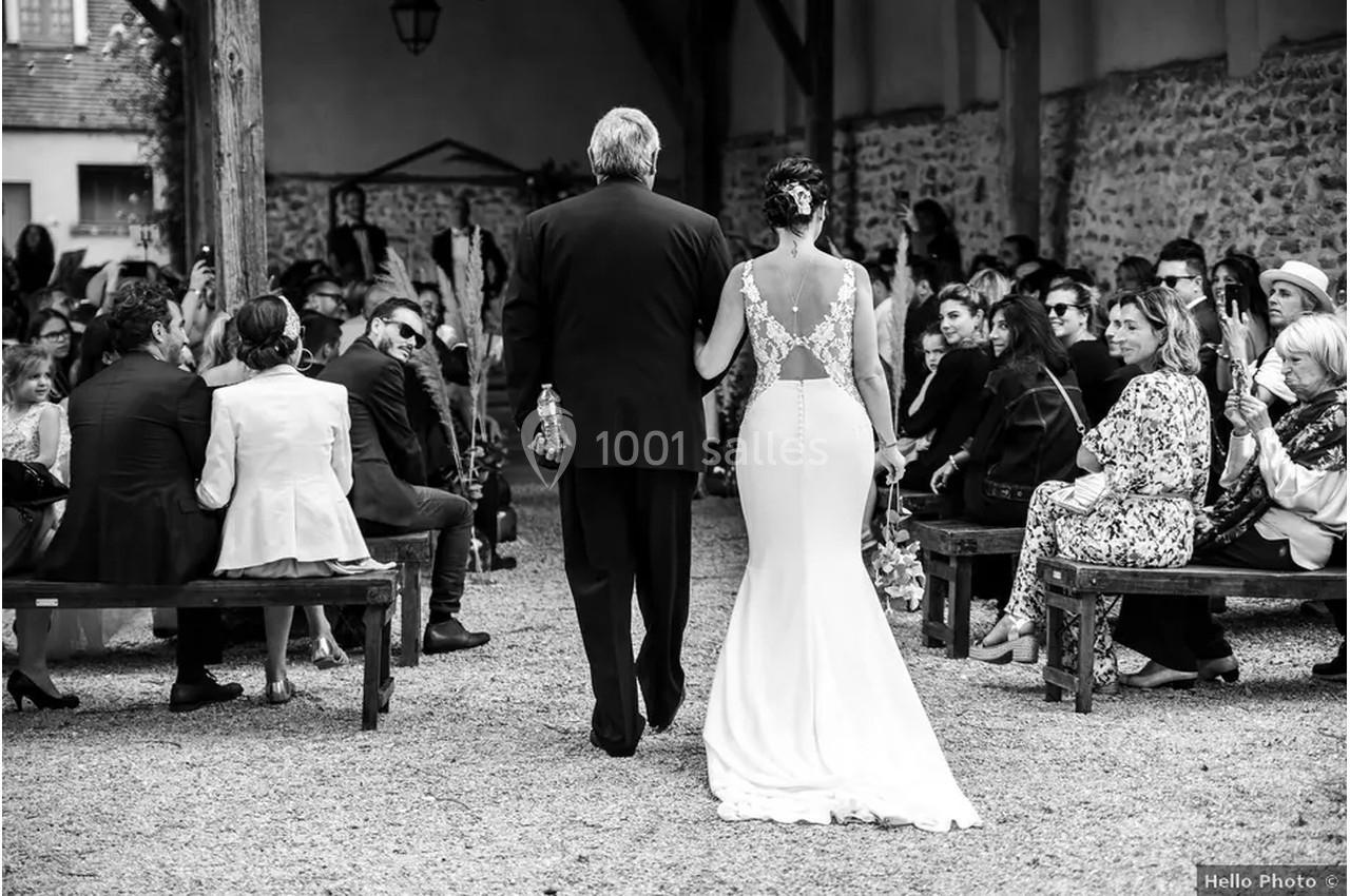 Un homme accompagne une mariée en robe blanche dans une allée, entourés d'invités assis lors d'une cérémonie en extérieur.