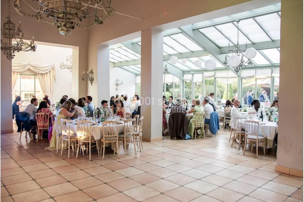 Salle lumineuse avec de grandes baies vitrées, décorée pour un événement, où des invités sont assis à des tables rondes.