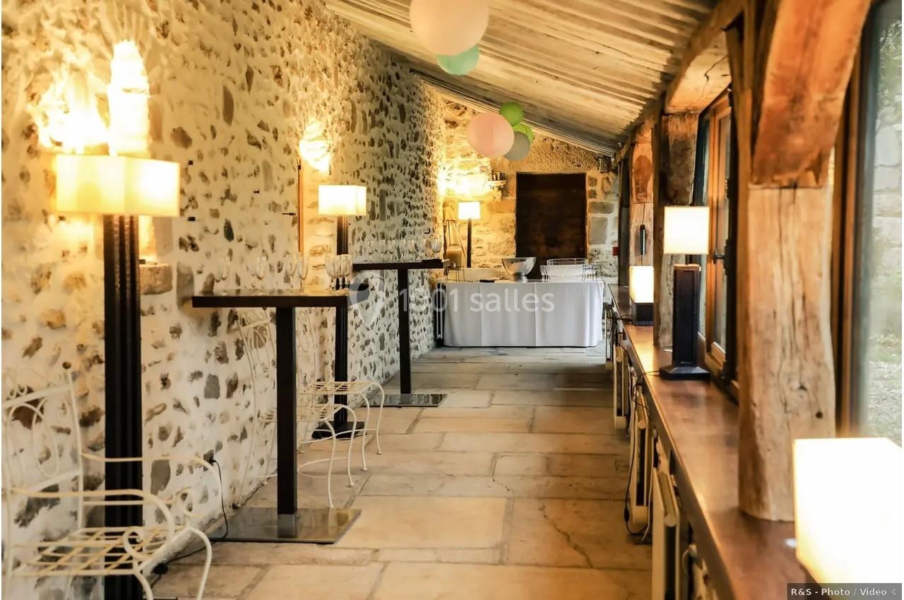 Salle en pierre avec tables hautes, chaises en métal, lampes allumées et une table dressée au fond.