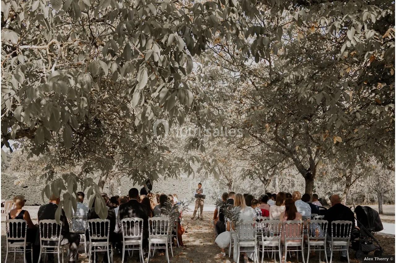 Cérémonie en plein air sous des arbres, avec des invités assis sur des chaises blanches face à un orateur.