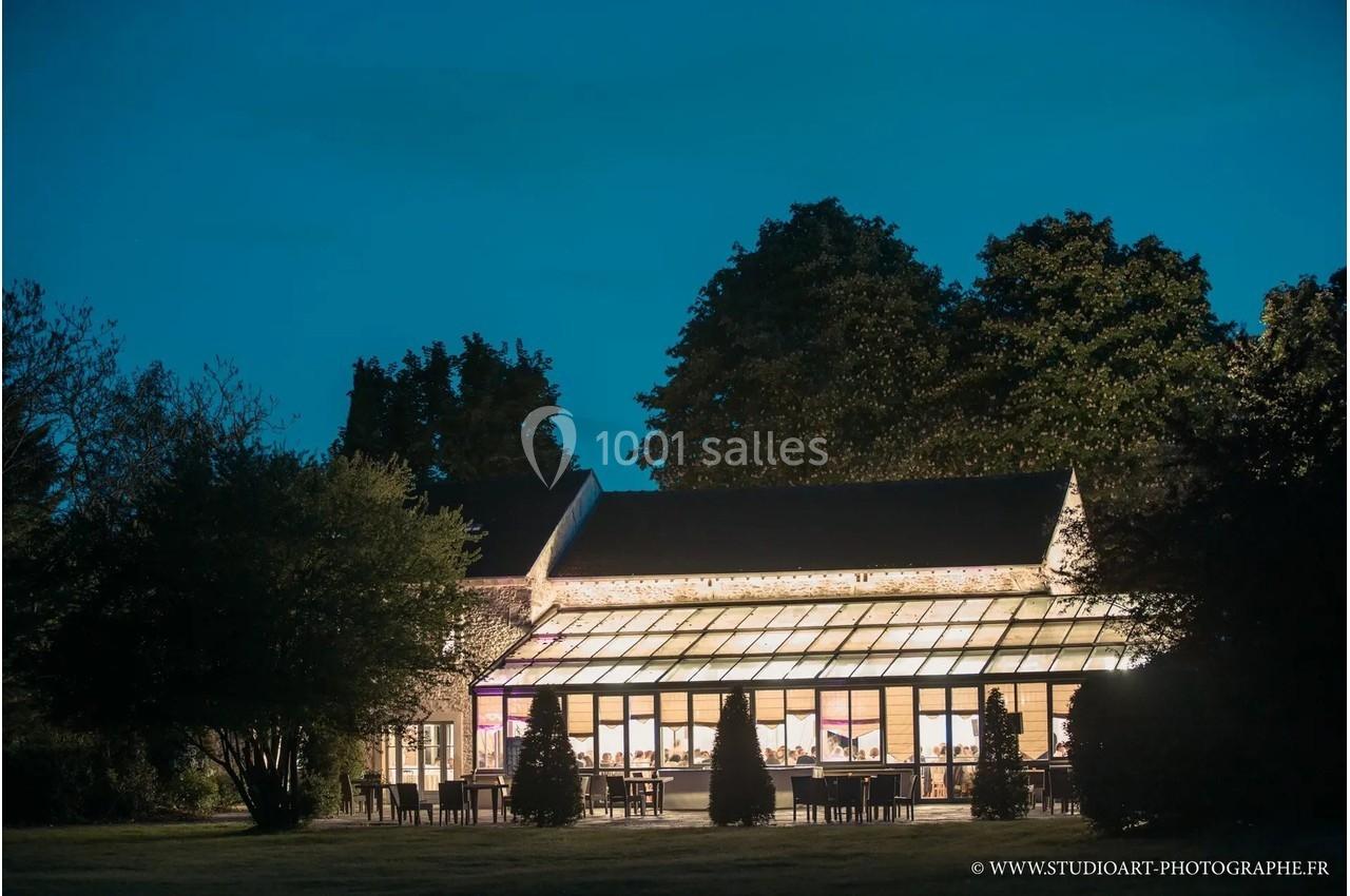 Bâtiment avec une grande verrière illuminée, entouré d'arbres, photographié à la tombée de la nuit.