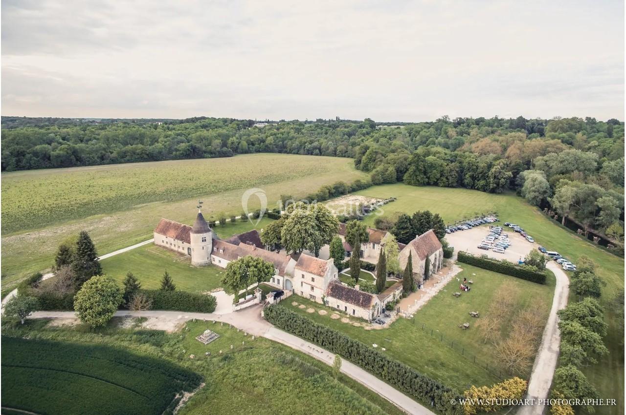 Vue aérienne d'un domaine rural avec bâtiments anciens, espaces verts et un parking entouré de champs et de forêt.