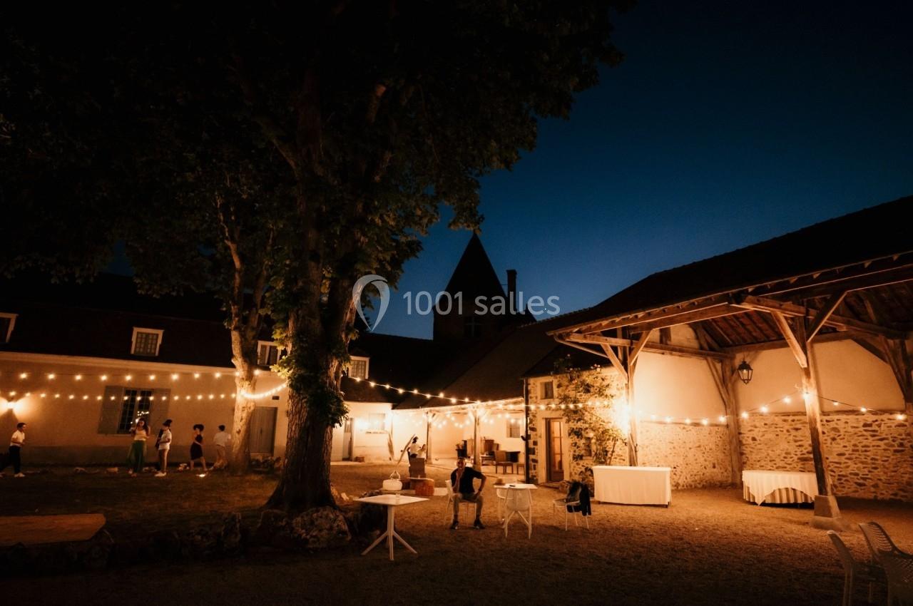 Cour extérieure éclairée par des guirlandes lumineuses au crépuscule, avec quelques personnes et bâtiments en arrière-plan.