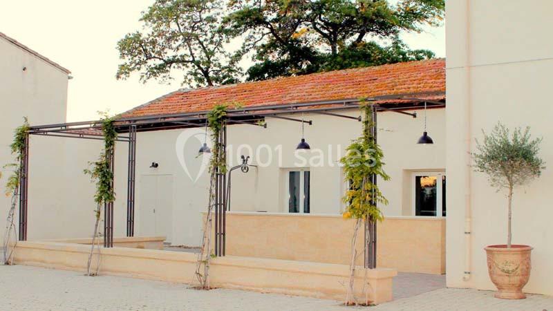 Façade d'un bâtiment clair avec terrasse couverte, plantes grimpantes et pots décoratifs, entourée d'arbres.